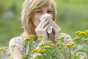 Woman blowing nose into tissue outside in spring, near yellow flowers
