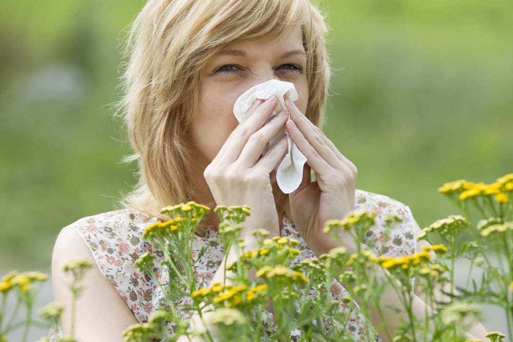 Woman blowing nose into tissue outside in spring, near yellow flowers