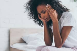 Young woman sitting on bed, hands on temples, looking distressed, migraine
